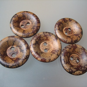 Może przedstawiać: Pięć brązowych i złotych, nakrapianych guzików z dwoma otworami każdy. Guziki są okrągłe i mają wypukły środek.