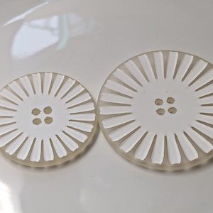 Könnte beinhalten: Zwei weiße Plastikknöpfe mit einem Sonnenstrahl-Design. Der größere Knopf hat einen Durchmesser von etwa 5 cm und der kleinere Knopf einen Durchmesser von etwa 3,8 cm.