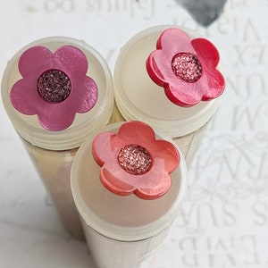 Op de afbeelding: Drie glazen potten met roze en rode bloemvormige deksels. De deksels hebben een glinsterend midden.