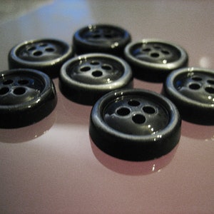 Könnte beinhalten: Eine Gruppe von sieben schwarzen Knöpfen mit jeweils vier Löchern. Die Knöpfe sind in einem Halbkreis auf einer hellrosa Oberfläche angeordnet.