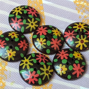 Könnte beinhalten: Fünf runde, dunkelbraune Knöpfe mit einem bemalten Blumenmuster aus gelben und korallenfarbenen Blüten, die mit kleinen grünen Blättern durchsetzt sind. Jeder Knopf hat zwei Löcher in der Mitte zur Befestigung und eine glatte Oberfläche.