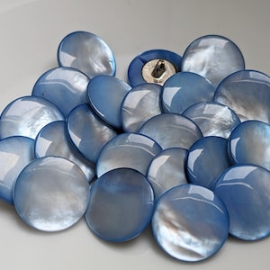 Può includere: Una collezione di 18 bottoni rotondi, blu e iridescenti con una finitura madreperlacea. I bottoni hanno un diametro di circa 2,5 cm.