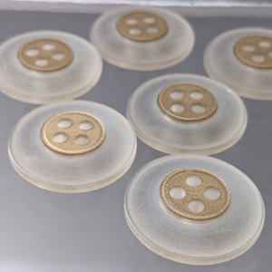Op de afbeelding: Zes doorzichtige plastic knoppen met gouden metalen centra, elk met vier gaten. De knoppen zijn in een halve cirkel op een grijs oppervlak gerangschikt.