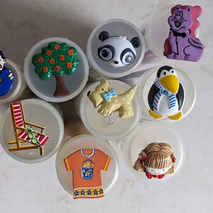Op de afbeelding: Assortiment van kleurrijke plastic knopen met verschillende ontwerpen, waaronder een panda, een boom, een pinguïn, een strandstoel, een meisje, een kat, een hond en een T-shirt.