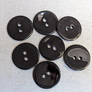 Op de afbeelding: Acht zwarte plastic knoppen met elk twee gaten. De knoppen zijn rond en hebben een glanzende afwerking.