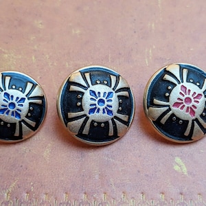 Può includere: Tre bottoni rotondi neri con accenti dorati e un design floreale stilizzato al centro. I fiori sono blu, rossi e rosa.