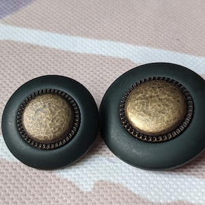 Può includere: Due pomelli rotondi, verde scuro, con un centro in ottone anticato e strutturato. I pomelli hanno un bordo decorativo e scanalato attorno al centro in ottone. Sono probabilmente per mobili o decorazioni per la casa.