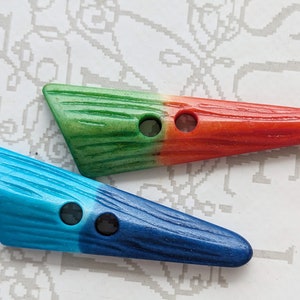 Könnte beinhalten: Zwei dekorative, längliche Knöpfe. Ein Knopf ist grün und rot, der andere blau. Beide Knöpfe haben eine Holzmaserung und zwei Löcher zum Annähen. Die Knöpfe liegen auf einem gemusterten Hintergrund.