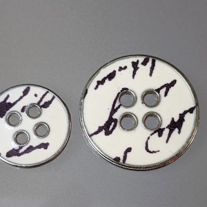 May include: Two white buttons with a silver rim. Both buttons have four holes and black ink markings. The larger button has the text "7-7-71" and "677" written on it. The smaller button has black ink markings that resemble abstract art.