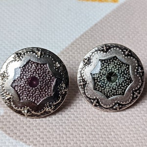 Op de afbeelding: Twee ronde, zilverkleurige knopen. Elke knoop heeft een decoratieve, bloemachtige rand. Eén knoop heeft een donkerpaars midden met een kleine, ronde, paarse edelsteen. De andere knoop heeft een donkergroen midden met een kleine, ronde, groene edelsteen.