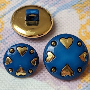 Puede incluir: Dos botones azules con corazones dorados y puntos dorados. El botón más grande tiene un diámetro de 2,5 cm. El botón más pequeño tiene un diámetro de 2 cm.