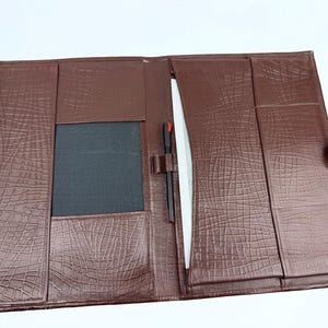 May include: Open brown leather portfolio with a crocodile skin texture. The interior features pockets, a pen holder with a black pen, and a white paper. A button closure secures the portfolio.