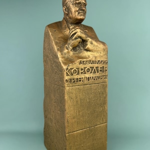 May include: A bronze sculpture of a man's bust, with hands clasped, mounted on a rectangular base. The base has engraved Cyrillic text. The sculpture has a weathered, aged look, with a golden-brown colour. The bust is a portrait of a notable person.