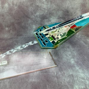 Pode incluir: Uma escultura decorativa em acrílico transparente de um avião estilizado. O avião apresenta um design turquesa, verde e branco com uma alça transparente e torcida e uma base transparente. O design do avião inclui uma paisagem com edifícios.