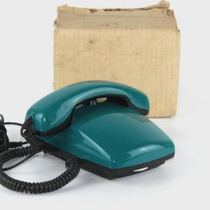May include: Teal vintage telephone with a black base and handset, coiled cord, and a cardboard box in the background. The phone has a sleek, retro design, suggesting a classic communication device.
