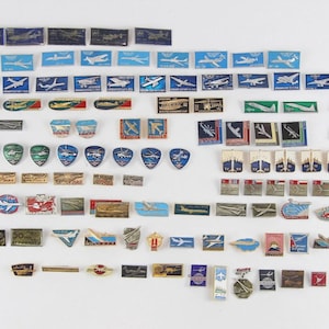 Op de afbeelding: Een verzameling vintage luchtvaartspelden en -badges, met verschillende vliegtuigontwerpen. De spelden zijn rechthoekig, schildvormig en cirkelvormig, met kleuren als blauw, groen, rood en goud. Sommige spelden hebben tekst.