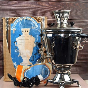 May include: A polished silver samovar with black handles and a tap, accompanied by its original box. The box features a blue and orange design with Cyrillic text. A black electrical cord is coiled in front.