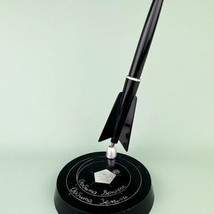Puede incluir: Un bolígrafo negro con punta dorada, montado sobre una base negra con texto. El bolígrafo está diseñado para parecerse a un cohete, con aletas y un acento plateado. La base tiene texto grabado y un diseño geométrico.