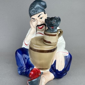 May include: A ceramic figurine of a seated man with a winking expression, wearing a dark hat, white shirt, and blue pants. He holds a small mug with a black creature on top. A tan barrel is in front of him.