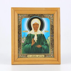 May include: A framed religious icon featuring a woman in a white head covering and green robe. The icon has a blue background with a black halo and gold accents. The frame is made of light brown wood.
