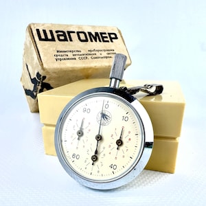 May include: A vintage stopwatch with a cream-colored face, featuring black numerals and three smaller dials. The stopwatch is encased in a silver metal frame and has a winding stem. A beige box with Cyrillic text is in the background.