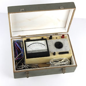 Puede incluir: Una caja de madera gris abierta que contiene un dispositivo de prueba eléctrica vintage. El dispositivo tiene un dial negro con marcas blancas, una perilla central y varios terminales de entrada. Se ven cables de diferentes colores dentro de la caja.