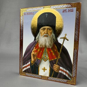 Op de afbeelding: Een rechthoekig religieus icoon met een portret van een bebaarde figuur in een zwarte monnikskap en paarse gewaden. De figuur houdt een gouden staf vast en maakt een zegenend gebaar. De rand is goudkleurig met ingewikkelde zilveren patronen.