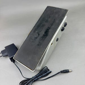 Può includere: Un pedale per effetti per chitarra color argento con un pedale nero. Il pedale ha tre manopole nere e un adattatore di alimentazione con un cavo nero. Il pedale è rettangolare e ha una superficie strutturata.