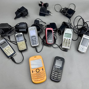 May include: A collection of various used cell phones in different colors, including black, silver, orange, and red. Several black power adapters with cords are also present. The phones have keypads and small screens, representing older mobile technology.