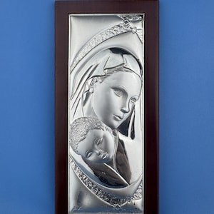 Puede incluir: Un relieve plateado de una madre y un niño, enmarcado en madera marrón oscuro. Las figuras están detalladas, sobre un fondo azul claro. La obra es una representación religiosa, ideal para la decoración.