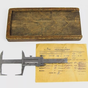Puede incluir: Un calibre metálico vintage, una caja de madera y un documento amarillento. El calibre es una herramienta de medición con una escala deslizante. La caja de madera es rectangular y tiene un aspecto desgastado. El documento tiene texto manuscrito.