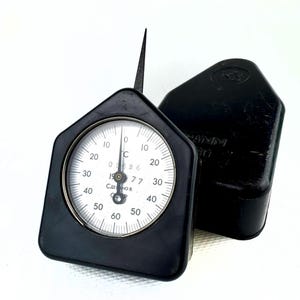 Könnte beinhalten: Ein schwarzer Messuhr mit Nadel und schwarzem Gehäuse. Das Zifferblatt hat weiße Zahlen und Markierungen mit dem Text "СДЕЛАНО В" und "190 77". Das Gehäuse hat eine Trapezform. Der Indikator wird zum Messen verwendet.