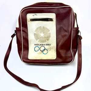 Könnte beinhalten: Burgunderfarbene Vintage-Umhängetasche mit weißer Paspelierung und langem Riemen. Die Tasche hat ein weißes Feld mit verblasstem Sternmuster, den olympischen Ringen und dem Text "MOCKBA-1980". Die Tasche hat eine Reißverschlusstasche und einen oberen Reißverschluss.