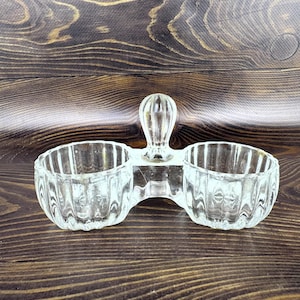 Può includere: Saliera doppia in vetro trasparente con manico. Le due ciotole sono collegate da una base in vetro trasparente. Il vetro ha un design a coste. La saliera è su una superficie di legno.