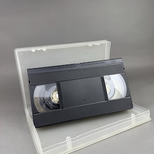 Puede incluir: Una cinta de vídeo VHS negra dentro de una caja de plástico transparente. La cinta muestra los carretes de la cinta y la caja está abierta. El texto "Do not touch the tape inside" es visible.