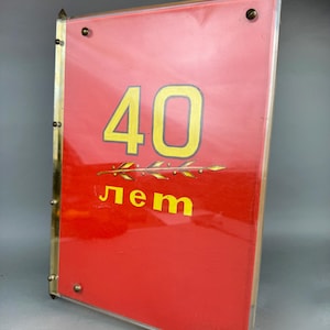 Puede incluir: Un letrero rectangular rojo con el número "40" en amarillo y la palabra "лет" en amarillo. El letrero tiene un marco dorado y está asegurado con tornillos. Probablemente sea un artículo conmemorativo.