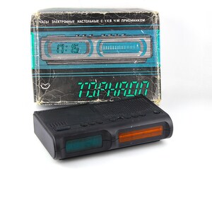 May include: A black digital alarm clock with a green and orange display, and a matching box. The clock displays the time 17:15 in green digits. The box has text in a foreign language and a teal and black design.