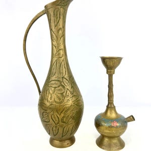May include: Two antique brass decorative items. A tall, slender pitcher with a curved handle and leaf pattern engraving. Next to it is a small, ornate brass object with a floral design and a spout. Both items have a vintage aesthetic.
