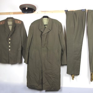 Op de afbeelding: Een olijfgroene militaire uniformset, bestaande uit een jasje met gouden knopen en epauletten, een lange jas, een broek en een pet. Het uniform wordt op hangers gepresenteerd. De pet heeft een rode band.
