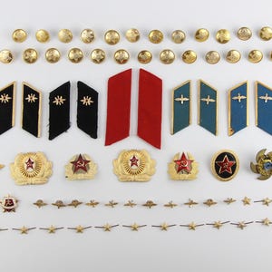 May include: A collection of vintage military insignia and buttons, including gold-colored buttons, shoulder boards in black, red, and blue, and various badges with stars and wings. The items are arranged on a white surface.