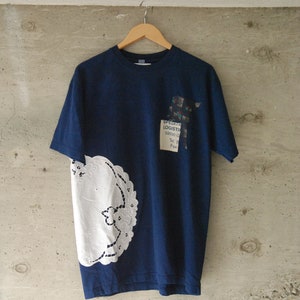 Può includere: Una t-shirt blu navy con un'applicazione di pizzo bianco sul lato sinistro. La t-shirt ha una piccola toppa sul lato destro con il testo "SPEDIZIONI LOGISTICHE 59950 G Tel. Fax"