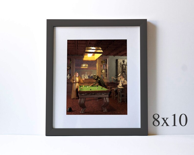 May include: A framed illustration of a pool room with two figures playing pool. The room is dimly lit with a single light fixture above the pool table. The pool table is green with white balls and a cue stick. The figures are dressed in casual clothing. The frame is dark gray with a white mat.