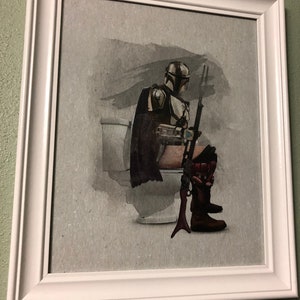May include: A framed print of a Star Wars character, The Mandalorian, sitting on a toilet. The character is wearing a helmet and armor, and is holding a rifle. The background is a watercolor wash of gray and black.