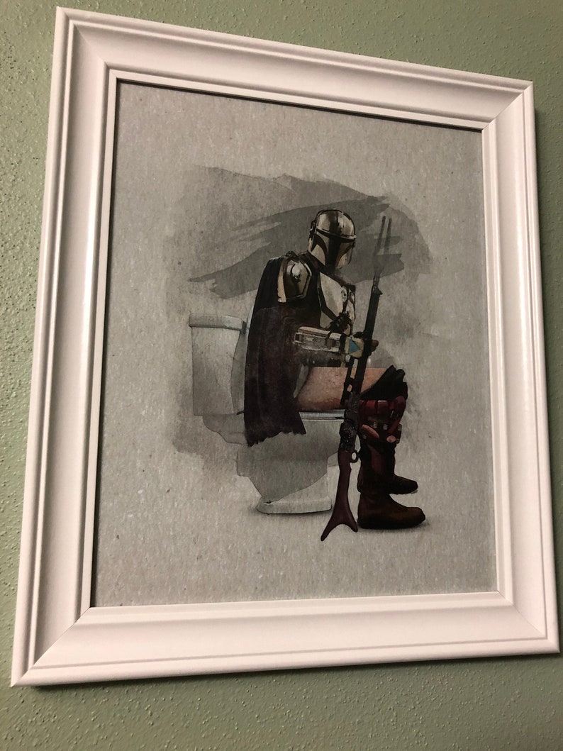 May include: A framed print of a Star Wars character, The Mandalorian, sitting on a toilet. The character is wearing a helmet and armor, and is holding a rifle. The background is a watercolor wash of gray and black.