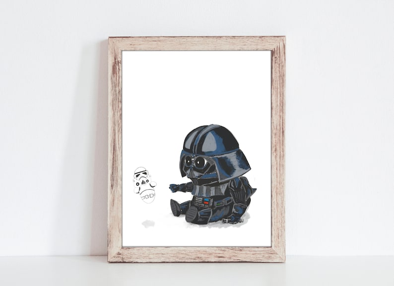 May include: A framed print featuring a cartoon illustration of a baby Darth Vader in a black and blue suit, helmet, and a small Stormtrooper. The artwork is set against a white background, with a light brown wooden frame.