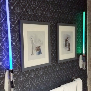 May include: Two framed pictures with Star Wars characters on a black and white wallpaper with a repeating pattern of Darth Vader and Stormtrooper helmets. Two light sabers are mounted on the wall, one blue and one green.