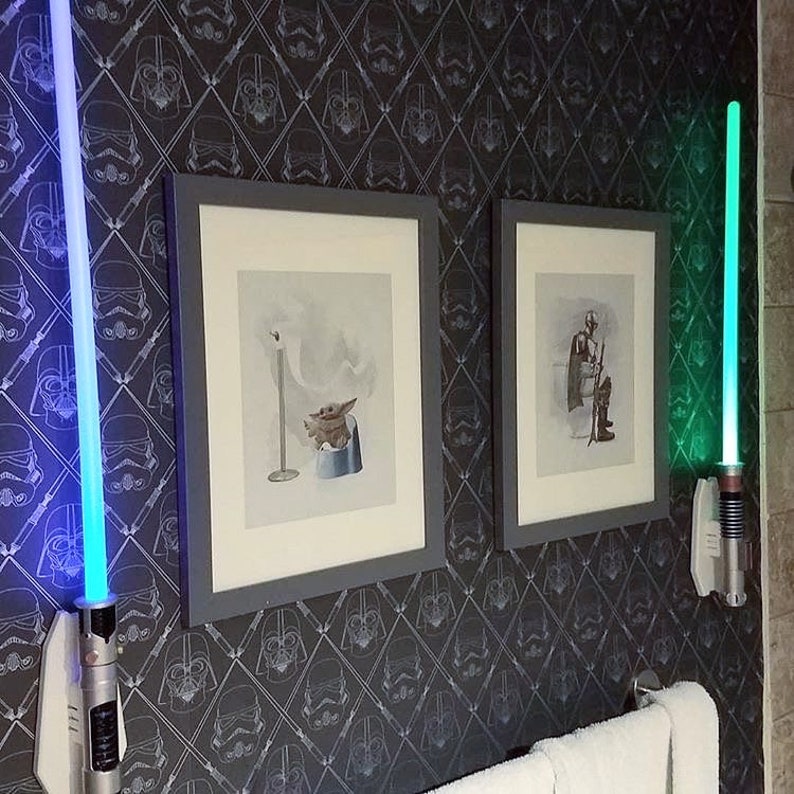 May include: Two framed pictures with Star Wars characters on a black and white wallpaper with a repeating pattern of Darth Vader and Stormtrooper helmets. Two light sabers are mounted on the wall, one blue and one green.
