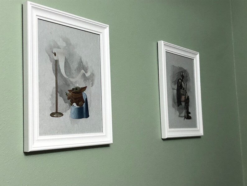 May include: Two framed prints on a green wall. The print on the left shows a cartoon character sitting on a toilet with a roll of toilet paper hanging above. The print on the right shows a cartoon character sitting on a toilet with a lightsaber.