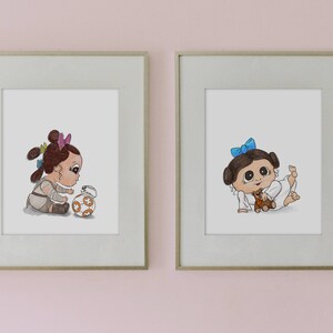May include: Two framed prints featuring cartoon illustrations of babies. One print shows a baby with brown hair and a BB-8 droid. The other print depicts a baby with a blue bow and a teddy bear. The prints are in light gray frames.