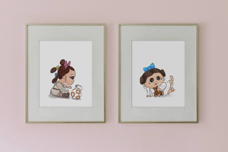 May include: Two framed prints featuring cartoon illustrations of babies. One print shows a baby with brown hair and a BB-8 droid. The other print depicts a baby with a blue bow and a teddy bear. The prints are in light gray frames.